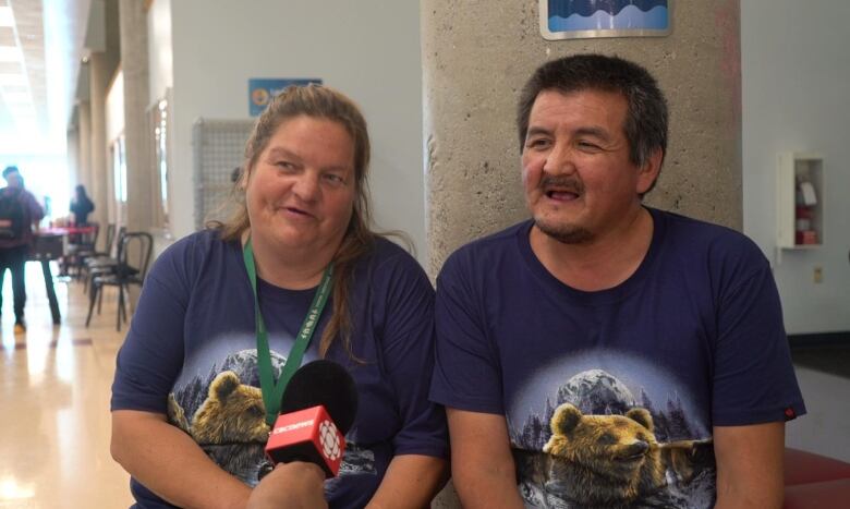 A man and a woman in matching t-shirts sit in a big facility talking into a CBC microphone.