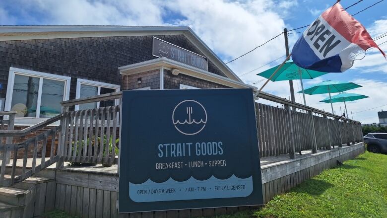 Building with grey cedar shingles, a large deck, an OPEN sign and a sign advertising the services and hours of the Strait Goods restaurant.