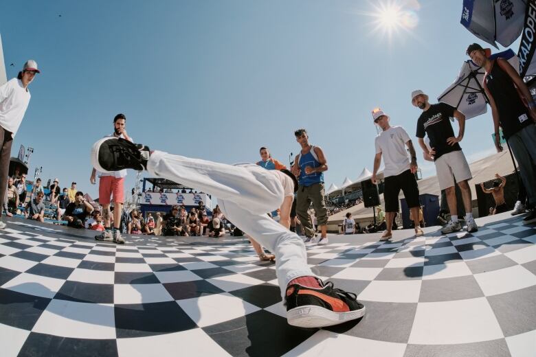 A man breakdancing on a checkered floor, several other men stand around him watching.