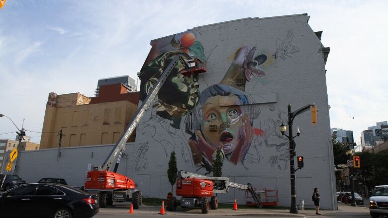 A big half-finished mural of two children. A man can be seen on a platform painting with spray paint.