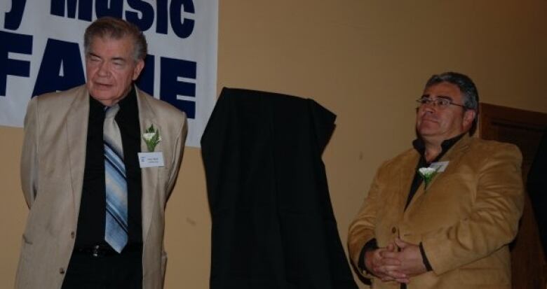 Two men in suits stand in a dark room, looking at something to the left of the camera. They both wear pale yellow flowers over their hearts.