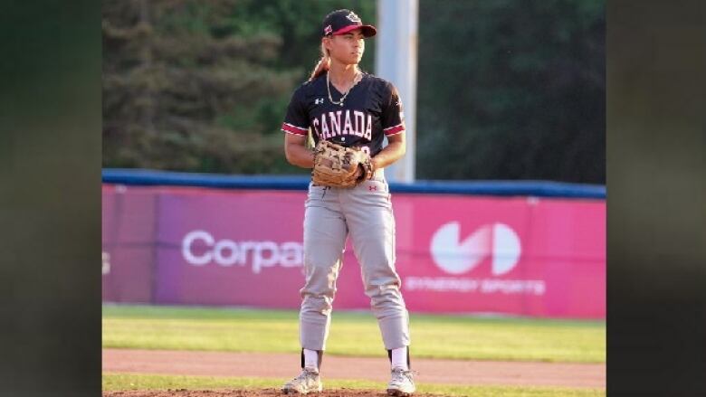 A woman wearing a baseball cap in a navy jersey that says Team Canada stands on a baseball field looking at home plate with her hand in her glove.