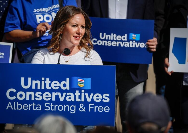 A woman wearing an off-white sweater is shown speaking behind a podium with a blue banner that reads: United Conservatives: Alberta Strong & Free.