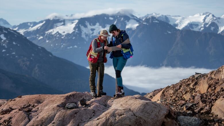 Two workers stand on top of a rock in a mountainous area of B.C., consulting a clipboard as excavation tools rest against their legs.