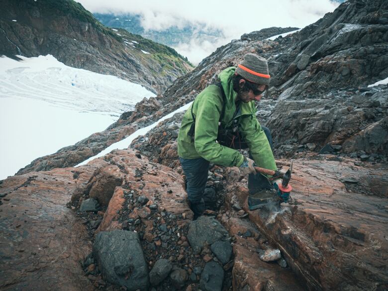 A man with a toque, sunglasses and green jacket uses an excavation tool to take rock samples on snow-covered mountainside.