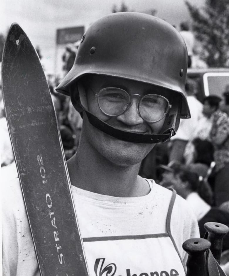 A person is shown in closeup, wearing a military-style helmet while holding skis and poles.