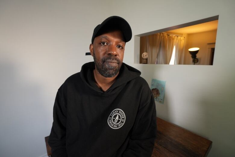 A man wearing a black hoodie and ballcap sits and looks at the camera. 