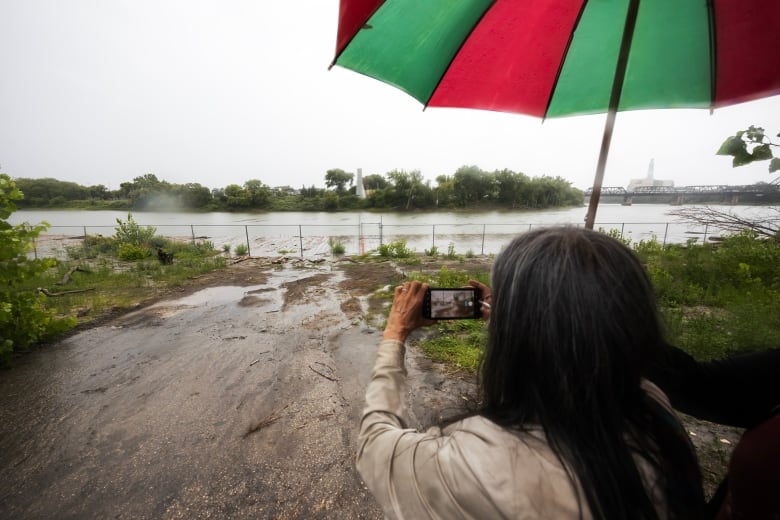 A person stands under an umbrella and takes a picture of a river with a cell phone.