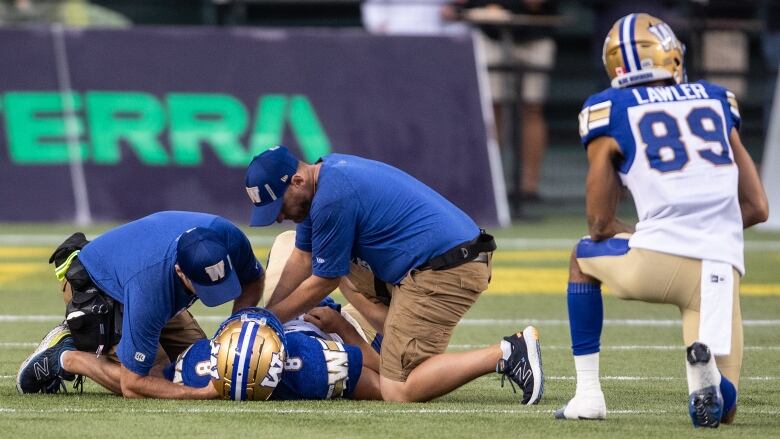 Two athletic trainers tend to a football player lying on his back, while a teammate kneels nearby.