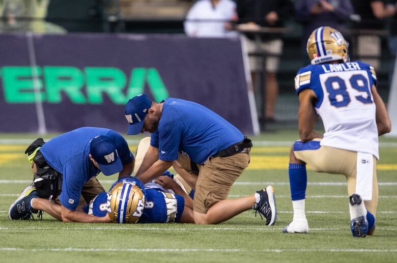 Two athletic trainers tend to a football player lying on his back, while a teammate kneels nearby.