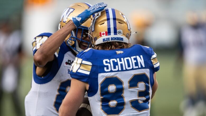 One football player smiles and taps a teammate on the top of his helmet.