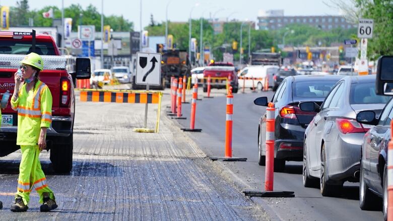 Two workers in bright yellow uniforms stand beside a line of cars on a stretch of road under construction.