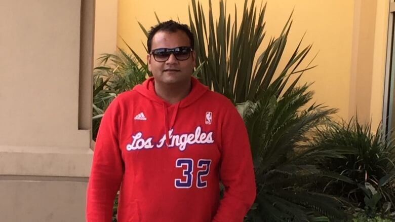 A man wearing sunglasses and a hoodie stands in front of some plants.