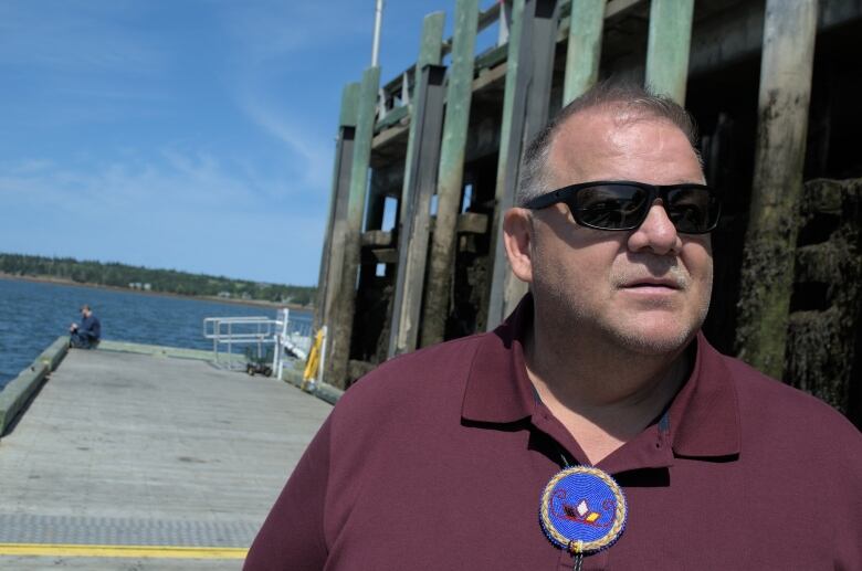 A man in sunglasses looks to the right of the camera. A man sits on the dock in the background, his fishing rod in the water.