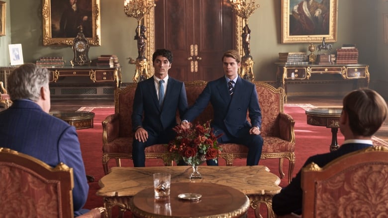 Two men sit on a velvet couch in an opulent room and hold hands.