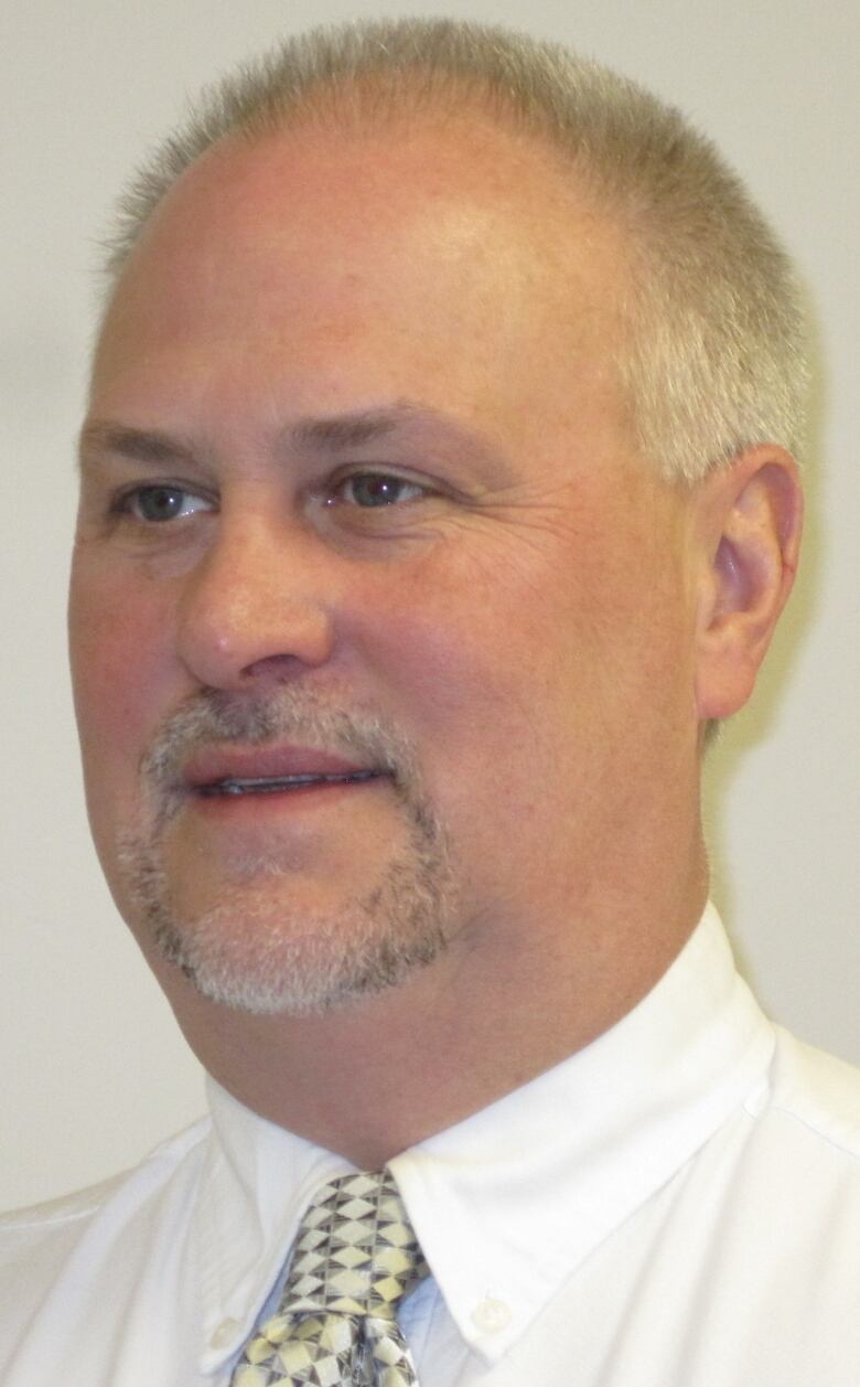 A head shot of a balding man with a grey moustache and goatee wearing a white dress shirt and tie.