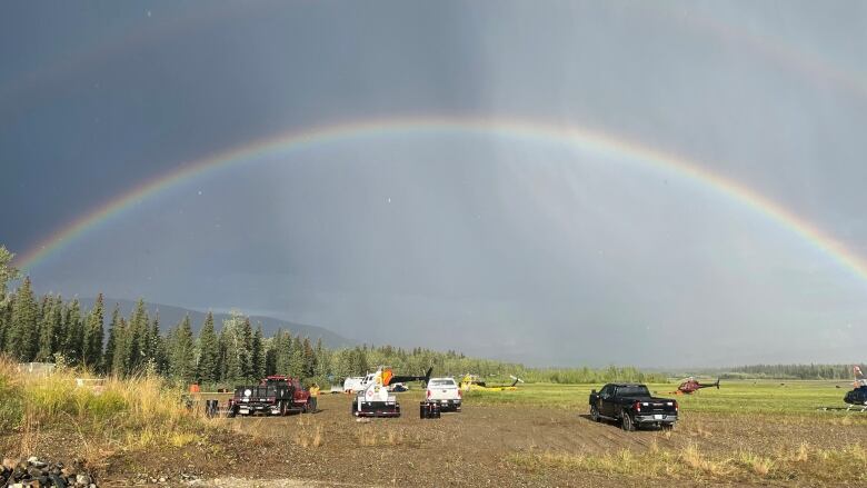 A rainbow is seen over an open field with vehicle and helicopters.