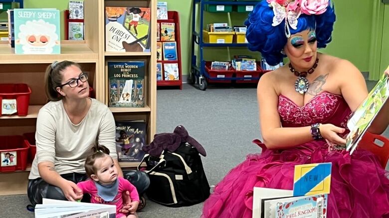 A drag queen is seen reading a story as a child and her mother sit on the floor and listen.