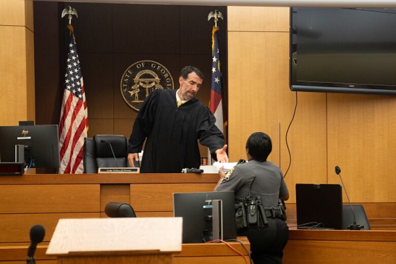 A man in a black robe behind a judge's bench is handed paper from a woman in uniform.