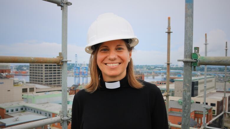 A young woman in a priest's collar and a hard hat stands on a rooftop cosntruction site. 