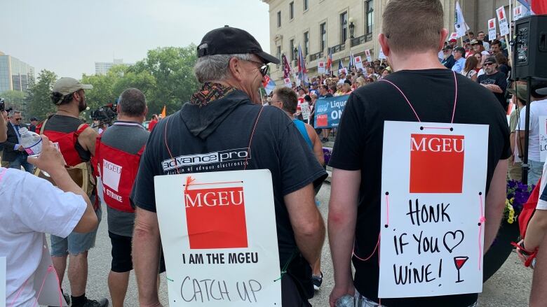 The back of two people with signs, one that that says 'catch up keep up' and another that says 'honk if you love wine' 