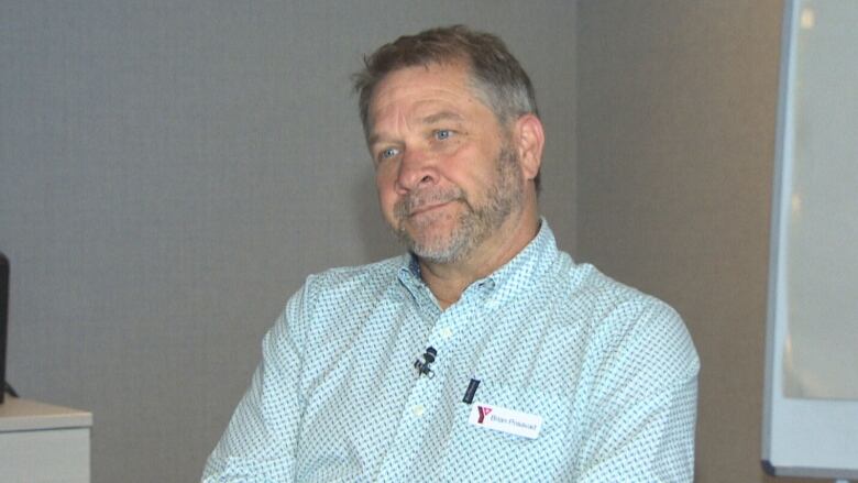 A man wearing a teal gingham shirt sits in front of a grey wall.