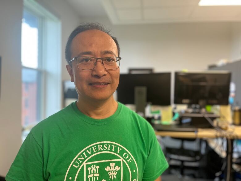 A man wears a green UPEI tshirt and stands in his office. A standing desk with two monitors is visible in the background.