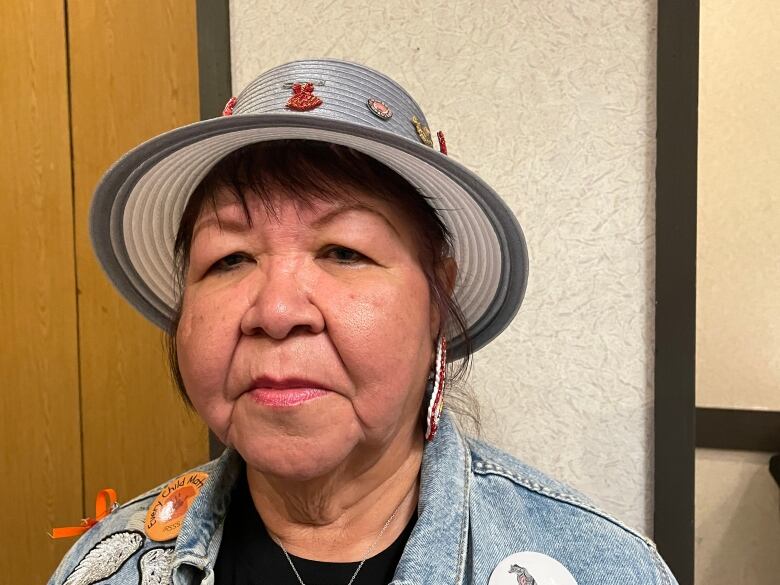 An Indigenous woman wearing a jean jacket, hat, traditional beaded earrings and a necklace looks sombre. 