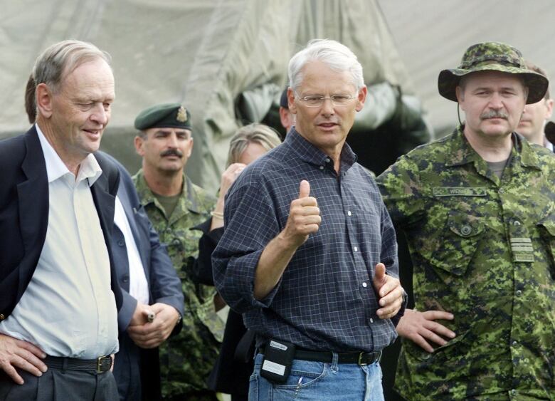 Two men stand together, flanked by army personnel. On with white hair gives the thumbs up.