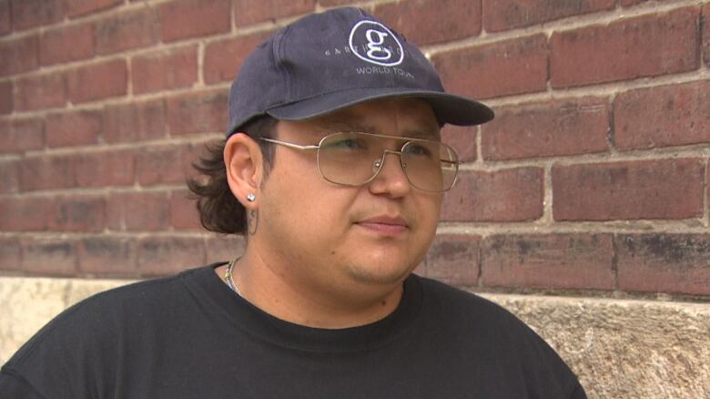 A person wearing aviator glasses, a black t-shirt and hat is seen looking forward.