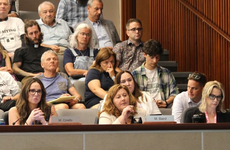 three women sit at desk in council chambers with audience members behind