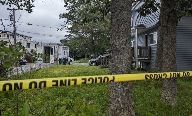 Police tape runs across the frame, with a blue house and emergency vehicle in the background.