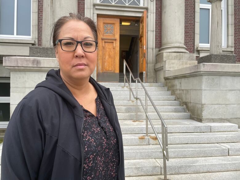 A middle aged woman with black hair wearing black-framed glasses and a black hoodie stands in front of a courthouse with a solemn look on her face.