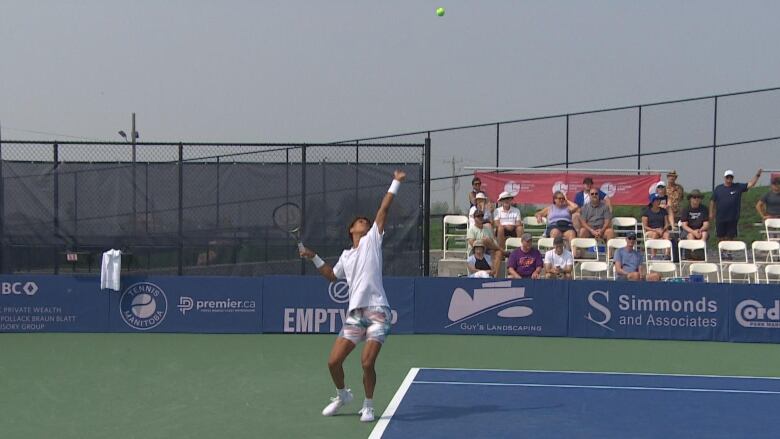 A player on a tennis court raises his racket toward a ball hanging in the air above him as spectators watch from the stands.