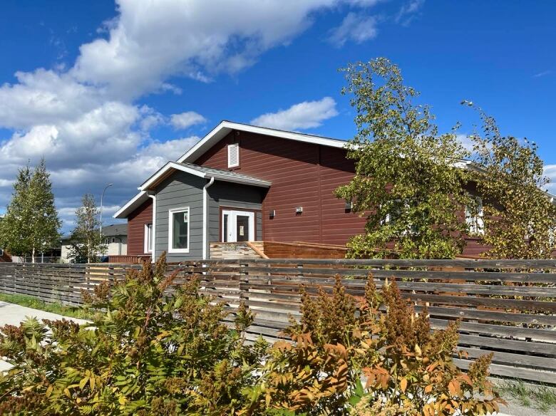 A small building sits behind a wooden fence and some bushes and trees.