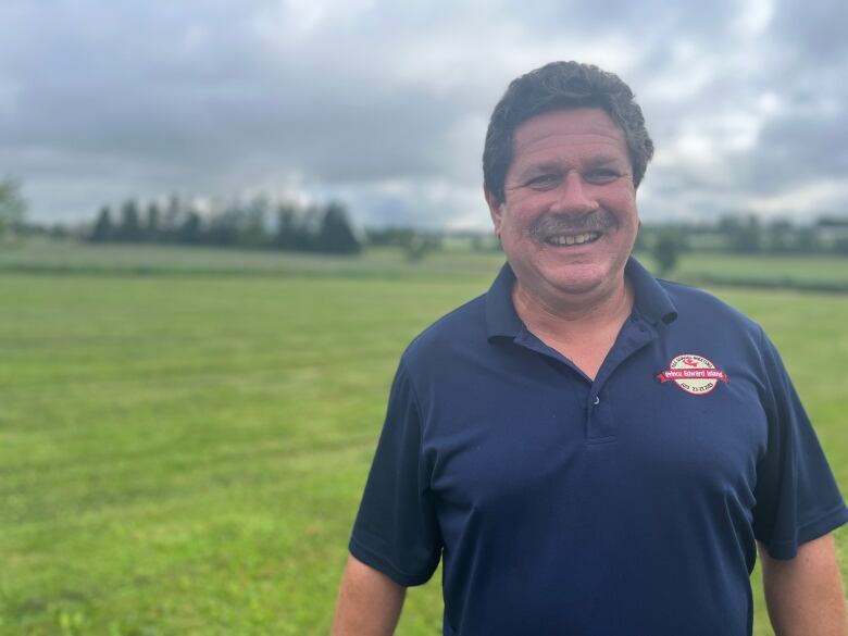 A man with dark hair and a mustache stands against a backdrop of lush green fields. He's wearing a blue polo shirt with an emblem.