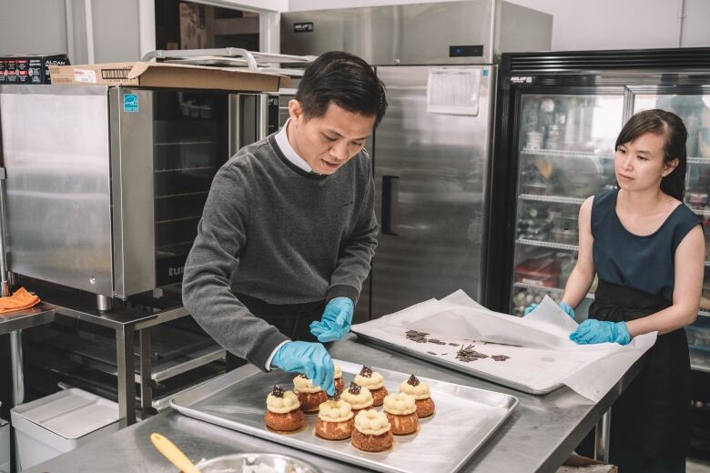 A man and a woman work at a metal table in a commercial kitchen, assembling delicate chouz pastries on a baking sheet.