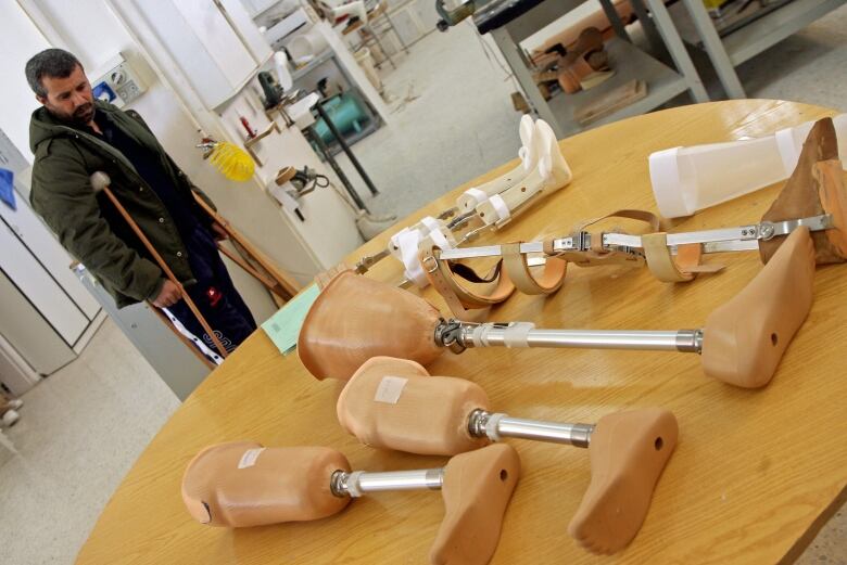 A man using crutches stands behind a table with several prosthetic legs on it. 