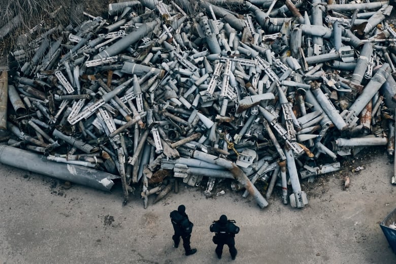 Two people stand in front of a large pile of spent rockets and bombs. 