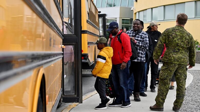 People board a school bus as a person in military garb watches on.