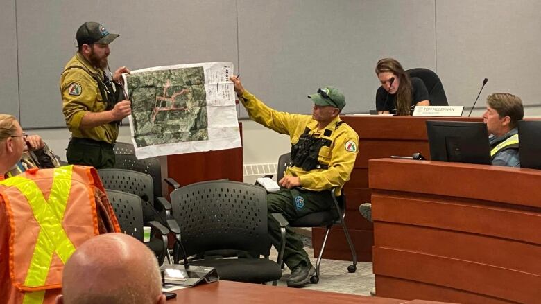 People sit in a meeting room talking while two wildland firefighters hold up a map.