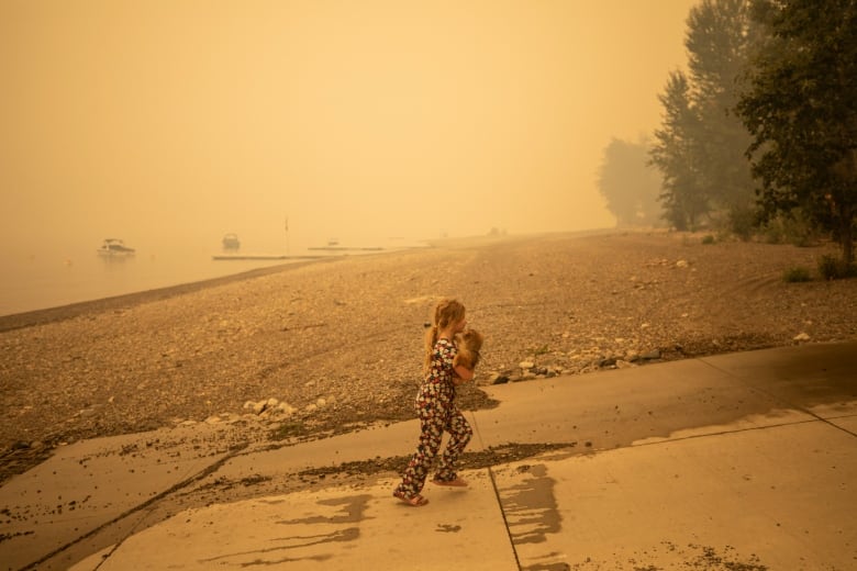 A girl wearing sandles carries a dog on her chest as she walks up from the water. The sky is hazy and orange around her.