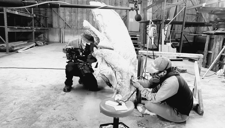 A man sits on the ground in a workshop, carving a giant piece of bone.