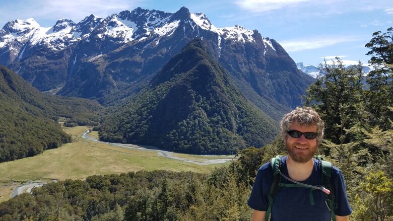 A man with curly grey hair and a large beard stands in front of a mountain vista with blue sky as he smiles for the camera.