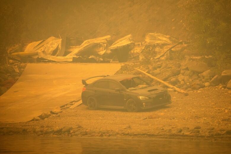 A black sports car, with the front window smashed, sits beside a lake, the air around it is brown and hazy, with fire-destroyed property behind it.