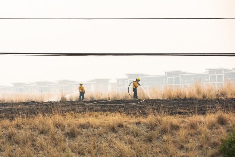 Firefighters mop up hot spots in a field in front of a row of houses on a hazy, possibly smoky day.