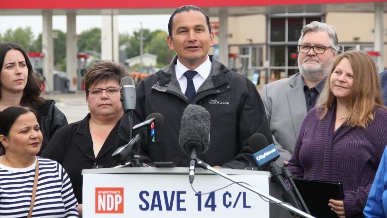 A man in a fall jacket stands at a podium, in front of a gas station across the street.