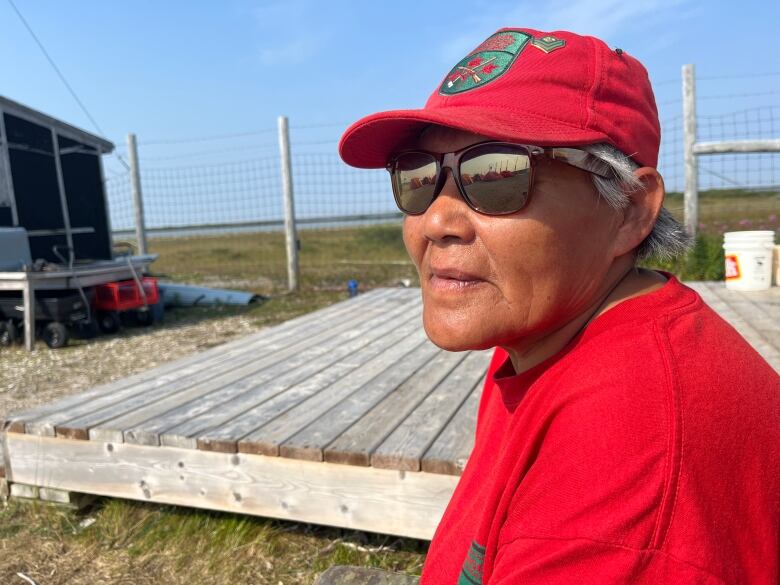 A woman wears a baseball cap, sweater and sunglasses.