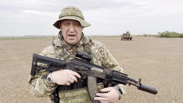 A man dressed in camouflaged fatigues and a bucket hat appears to be talking as he standings in the desert holding a large gun. A truck is driving away in the background.