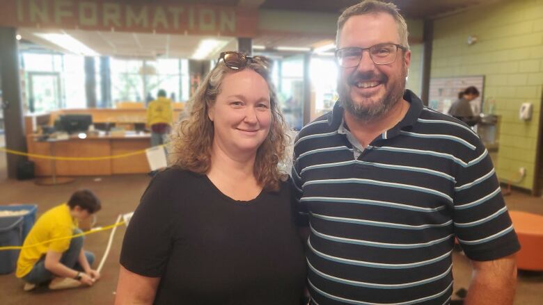 A man and a woman smile at the camera. In the background, their daughter builds Lego on the floor of the library.